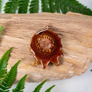May include: A brown resin pendant with a mandala design and a clear sunstone in the center. The pendant is on a piece of wood with green ferns.