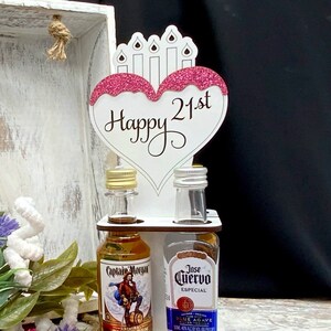 May include: A white wooden stand with two miniature bottles of liquor. The bottles are labeled "Captain Morgan Spiced Rum" and "Jose Cuervo Especial Blue Agave". A pink and white heart-shaped sign with the text "Happy 21st" is attached to the top of the stand.