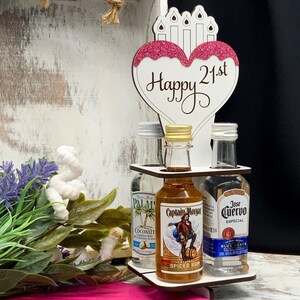 May include: A wooden stand with a pink heart-shaped sign that says "Happy 21st". The stand holds three miniature bottles of liquor: Captain Morgan Spiced Rum, Jose Cuervo Especial Tequila, and Coconut Rum.