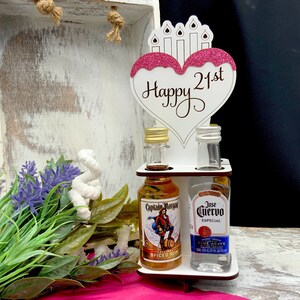 May include: A white wooden stand with a pink heart-shaped sign that says "Happy 21st" with candles on top. The stand holds two miniature bottles of liquor, one with Captain Morgan Spiced Rum and the other with Jose Cuervo Especial Blue Agave Tequila.