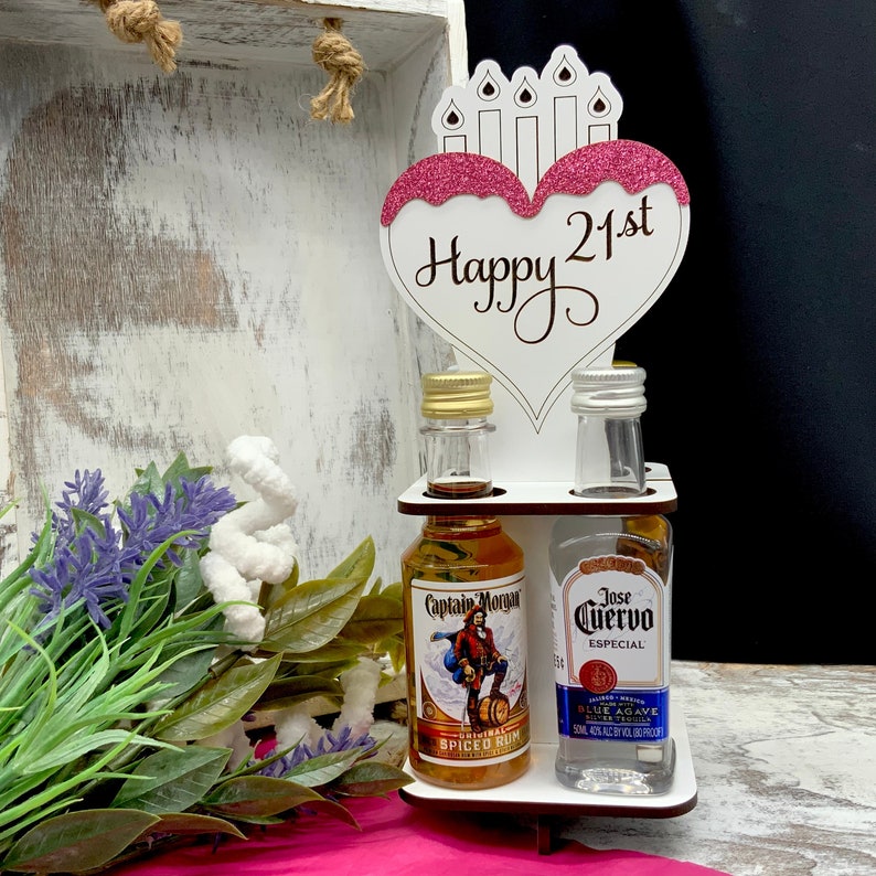 May include: A white wooden stand with a pink heart cutout that says "Happy 21st" with pink glitter. The stand has two shelves for holding two miniature bottles of liquor. One bottle is Captain Morgan Spiced Rum and the other is Jose Cuervo Especial Blue Agave Tequila.