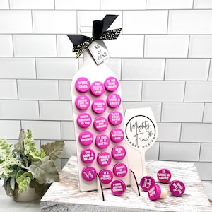 Könnte beinhalten: Ein weißer, holzerner Wein-Flaschenständer mit rosa und weißen Weinflaschenverschlüssen. Die Verschlüsse haben verschiedene Sprüche darauf, wie "Wine Diva", "My Blood Type is Wine", "Therapy in a Bottle", "Wine is a Carb?", "Let's Drink About It", "To the Birthday Babe", "Sip Back & Relax", "Mama's Sippy Cup", "It's Wine O'Clock", "8-00s are for Drinking", "Wine Will Fix It", and "Wine Diva".