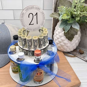 May include: A white wooden tiered cake stand with a blue ribbon and a blue tulle skirt. The stand is filled with miniature liquor bottles and rolled up dollar bills. A white circle with the number 21 and the text "Happy Birthday" is on top of the stand.