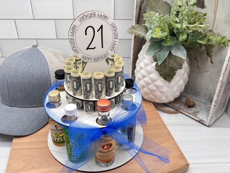 May include: A white wooden tiered cake stand with a blue ribbon and a blue tulle skirt. The stand is filled with miniature liquor bottles and rolled up dollar bills. A white circle with the number 21 and the text "Happy Birthday" is on top of the stand.