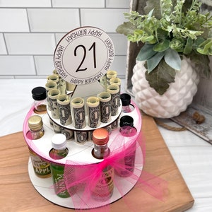 May include: A two-tiered cake stand with a pink tulle bow and a round topper that says "Happy Birthday!" with the number "21" in the center. The stand is filled with small liquor bottles and rolled up dollar bills.