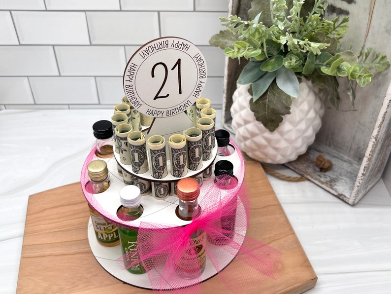 May include: A two-tiered cake stand with a pink tulle bow and a round topper that says "Happy Birthday!" with the number "21" in the center. The stand is filled with small liquor bottles and rolled up dollar bills.