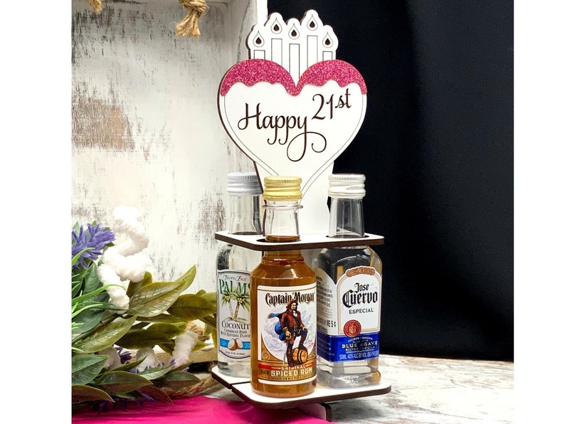 May include: A wooden liquor bottle holder with a pink heart-shaped sign that says "Happy 21st". The holder has three slots for mini bottles of liquor. The bottles are labeled "Captain Morgan", "Jose Cuervo", and "Palm Coconut".