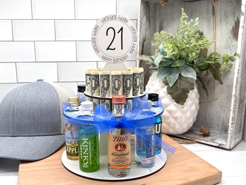 May include: A two-tiered cake stand with a blue ribbon and a sign that says "Happy Birthday! 21 Happy Birthday!"  The stand is filled with miniature liquor bottles and rolled up dollar bills.