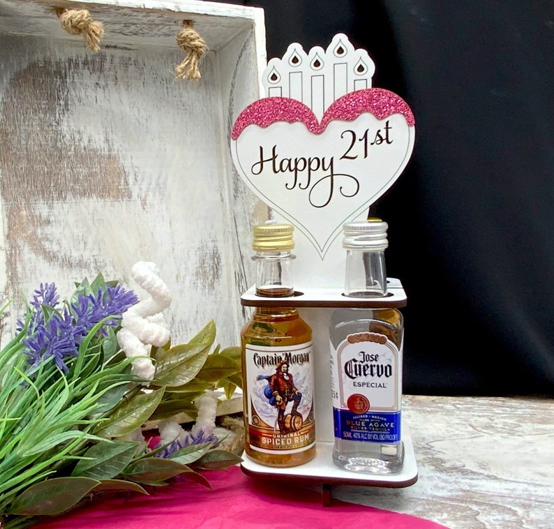 May include: A white wooden stand with two miniature bottles of liquor. The bottles are labeled "Captain Morgan Spiced Rum" and "Jose Cuervo Especial Blue Agave". A pink and white heart-shaped sign with the text "Happy 21st" is attached to the top of the stand.