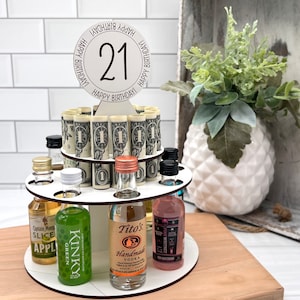 May include: A white wooden tiered stand with 21 rolled up dollar bills and miniature liquor bottles. The top tier has a white circle with the number 21 and the words "Happy Birthday!" written around it.