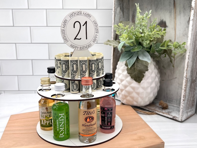 May include: A white wooden tiered stand with 21 rolled up dollar bills and miniature liquor bottles. The top tier has a white circle with the number 21 and the words "Happy Birthday!" written around it.