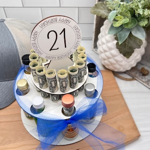 May include: A white wooden tiered cake stand with a blue ribbon and a sign that says "Happy Birthday" with the number 21 in the center. The stand is filled with miniature liquor bottles and rolled up dollar bills.