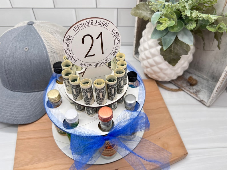 May include: A white wooden tiered cake stand with a blue ribbon and a sign that says "Happy Birthday" with the number 21 in the center. The stand is filled with miniature liquor bottles and rolled up dollar bills.