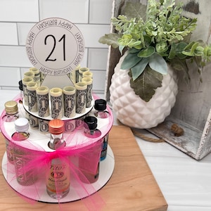 May include: A pink and white tiered cake stand with a pink bow and a clear bottle of Tito's Handmade Vodka. The stand is decorated with miniature liquor bottles and rolled up dollar bills. A white circle with the number 21 and the words "Happy Birthday" is on top of the stand.