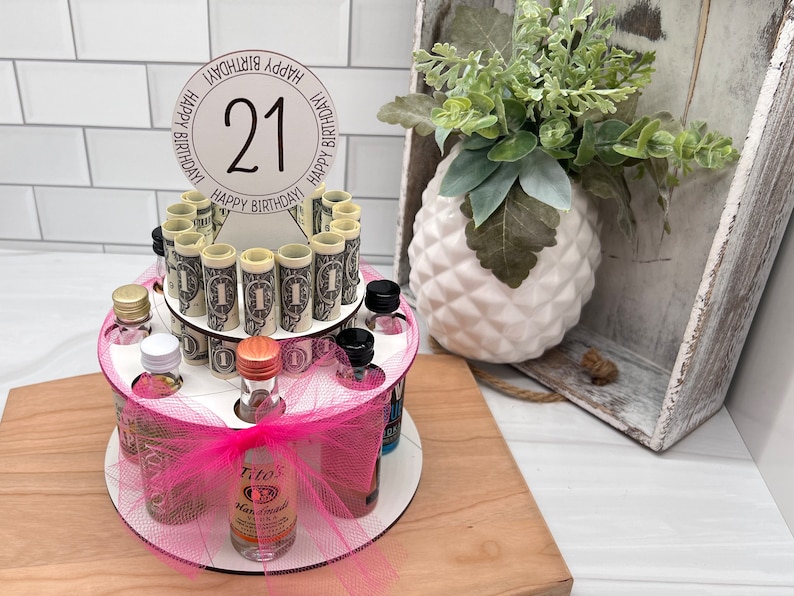 May include: A pink and white tiered cake stand with a pink bow and a clear bottle of Tito's Handmade Vodka. The stand is decorated with miniature liquor bottles and rolled up dollar bills. A white circle with the number 21 and the words "Happy Birthday" is on top of the stand.
