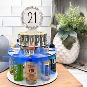 May include: A 21st birthday cake made of mini liquor bottles and rolled up dollar bills. The cake is decorated with a blue ribbon and a white sign that says "Happy Birthday 21 Happy Birthday".