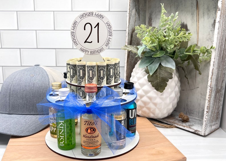 May include: A 21st birthday cake made of mini liquor bottles and rolled up dollar bills. The cake is decorated with a blue ribbon and a white sign that says "Happy Birthday 21 Happy Birthday".