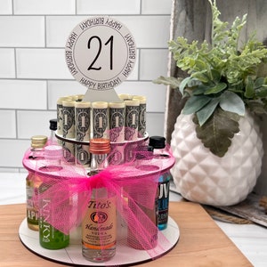 May include: A two-tiered birthday cake made of rolled dollar bills and miniature liquor bottles. The cake is decorated with a pink bow and a sign that says "Happy Birthday! 21". The cake sits on a wooden surface.