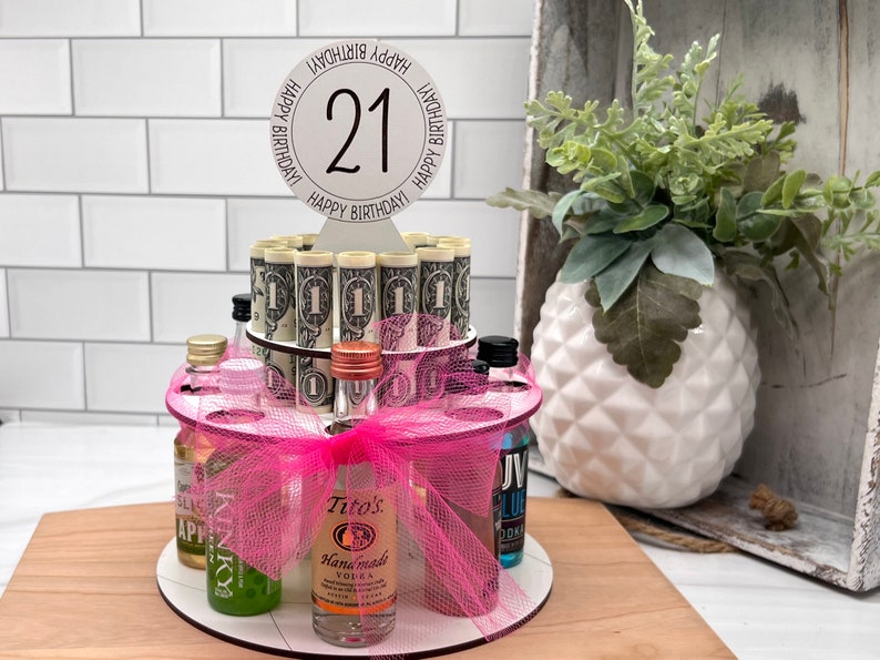 May include: A two-tiered birthday cake made of rolled dollar bills and miniature liquor bottles. The cake is decorated with a pink bow and a sign that says "Happy Birthday! 21". The cake sits on a wooden surface.