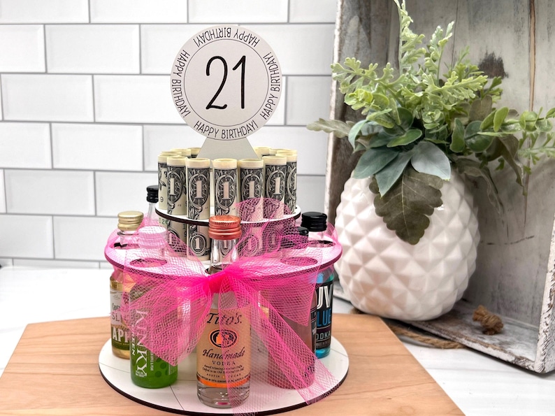 May include: A pink and white tiered cake with a round white topper that says "Happy Birthday! 21 Happy Birthday!" The cake is decorated with rolled up dollar bills and miniature liquor bottles.