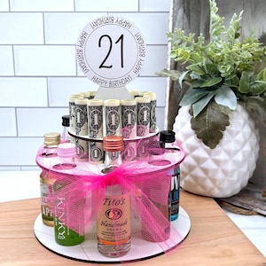 May include: A pink and white tiered cake stand with a pink ribbon and a white circle with the number 21 and the text "Happy Birthday" around it. The stand is filled with mini liquor bottles and rolled up dollar bills.