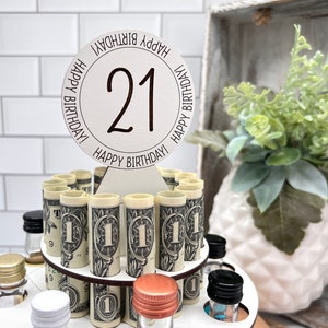 May include: A wooden tiered stand with a round white sign that says "Happy Birthday! 21 Happy Birthday!" The stand is filled with rolled up dollar bills and small glass bottles with various colored caps.