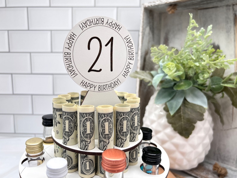 May include: A wooden tiered stand with a round white sign that says "Happy Birthday! 21 Happy Birthday!" The stand is filled with rolled up dollar bills and small glass bottles with various colored caps.