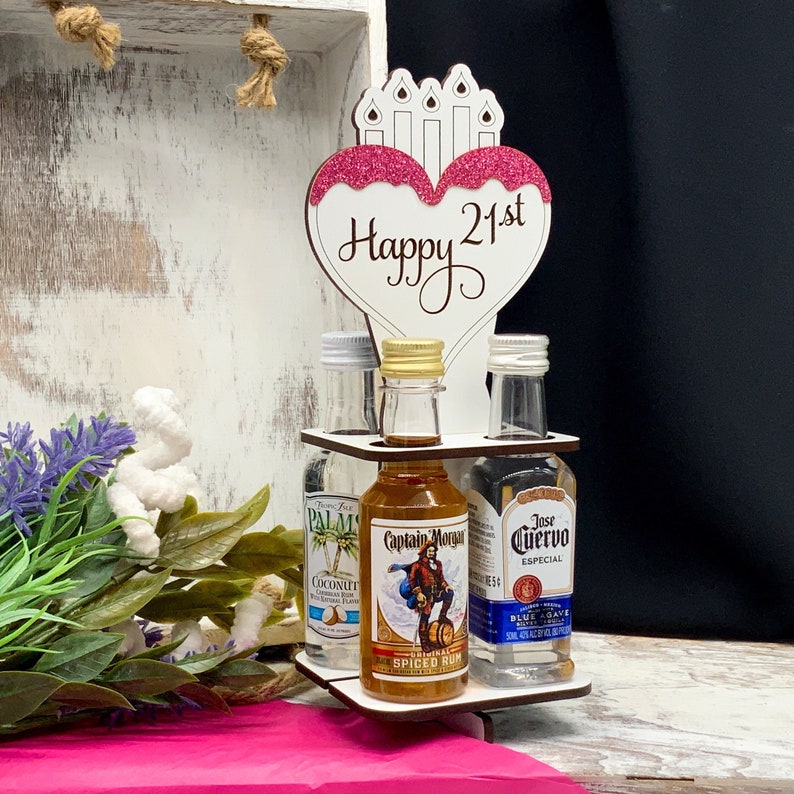 May include: Wooden liquor bottle holder with a pink heart shaped sign that says "Happy 21st". The holder has four slots for mini liquor bottles. The bottles are labeled "Captain Morgan", "Jose Cuervo", and "Palm Coconut".