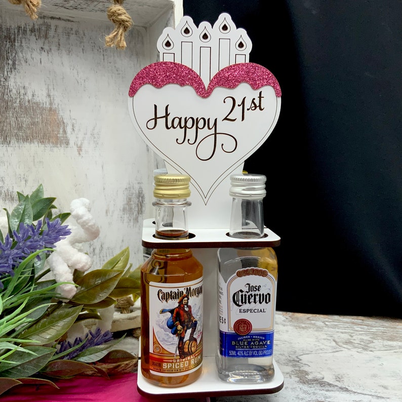 May include: White wooden stand with a heart-shaped sign that says "Happy 21st" with pink glitter accents. The stand holds two miniature bottles of alcohol, one with Captain Morgan Spiced Rum and the other with Jose Cuervo Especial Blue Agave Silver Tequila.