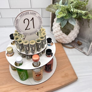 May include: A white tiered cake stand with a round wooden base. The stand is decorated with rolled up dollar bills and miniature liquor bottles. The top tier has a white circle with the number 21 and the text "Happy Birthday" repeated around the circle.