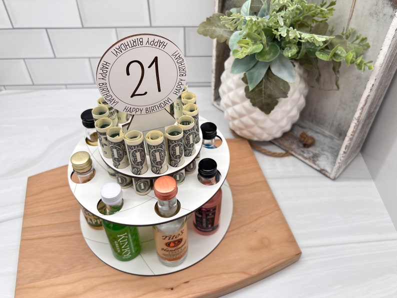 May include: A white tiered cake stand with a round wooden base. The stand is decorated with rolled up dollar bills and miniature liquor bottles. The top tier has a white circle with the number 21 and the text "Happy Birthday" repeated around the circle.