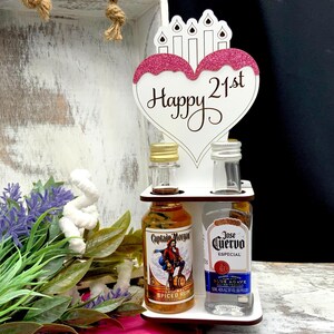 May include: A white wooden stand with a heart-shaped sign that says "Happy 21st" with pink glitter. The stand has two shelves for holding two miniature bottles of liquor. One bottle is Captain Morgan Spiced Rum and the other is Jose Cuervo Especial Blue Agave Silver Tequila.
