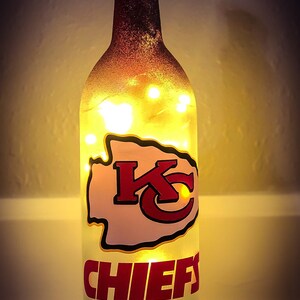 May include: A clear glass wine bottle with a frosted finish and a red, white, and yellow Kansas City Chiefs logo. The bottle is lit from within with white fairy lights.