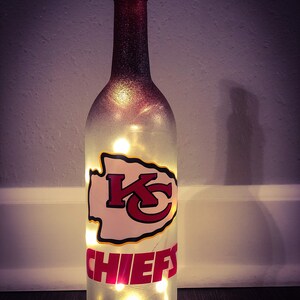 May include: A clear glass wine bottle with a red and white Kansas City Chiefs logo and the word "CHIEFS" on the front. The bottle is lit from inside with white fairy lights.