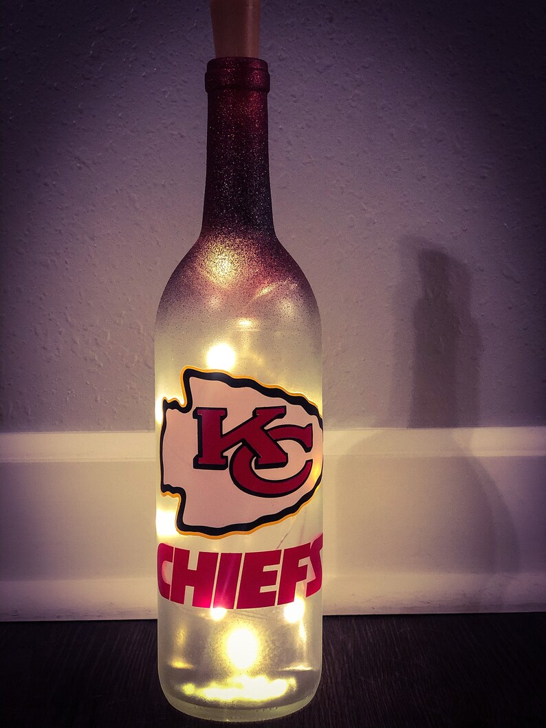 May include: A clear glass wine bottle with a red and white Kansas City Chiefs logo and the word "CHIEFS" on the front. The bottle is lit from inside with white fairy lights.