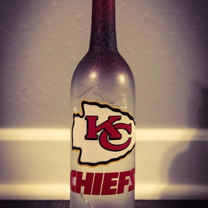 May include: A clear glass bottle with a red and black glitter finish. The bottle has a cork stopper and a white decal with the Kansas City Chiefs logo and the word "CHIEFS" in red letters.