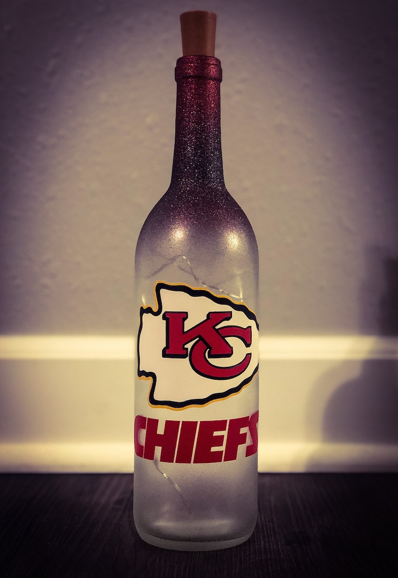 May include: A clear glass bottle with a red and black glitter finish. The bottle has a cork stopper and a white decal with the Kansas City Chiefs logo and the word "CHIEFS" in red letters.