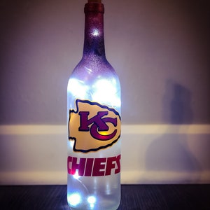 May include: A clear glass bottle with a white and purple ombre paint finish. The bottle has a cork stopper and features a red and yellow Kansas City Chiefs logo with the word "CHIEFS" in red lettering. The bottle is lit with white fairy lights.