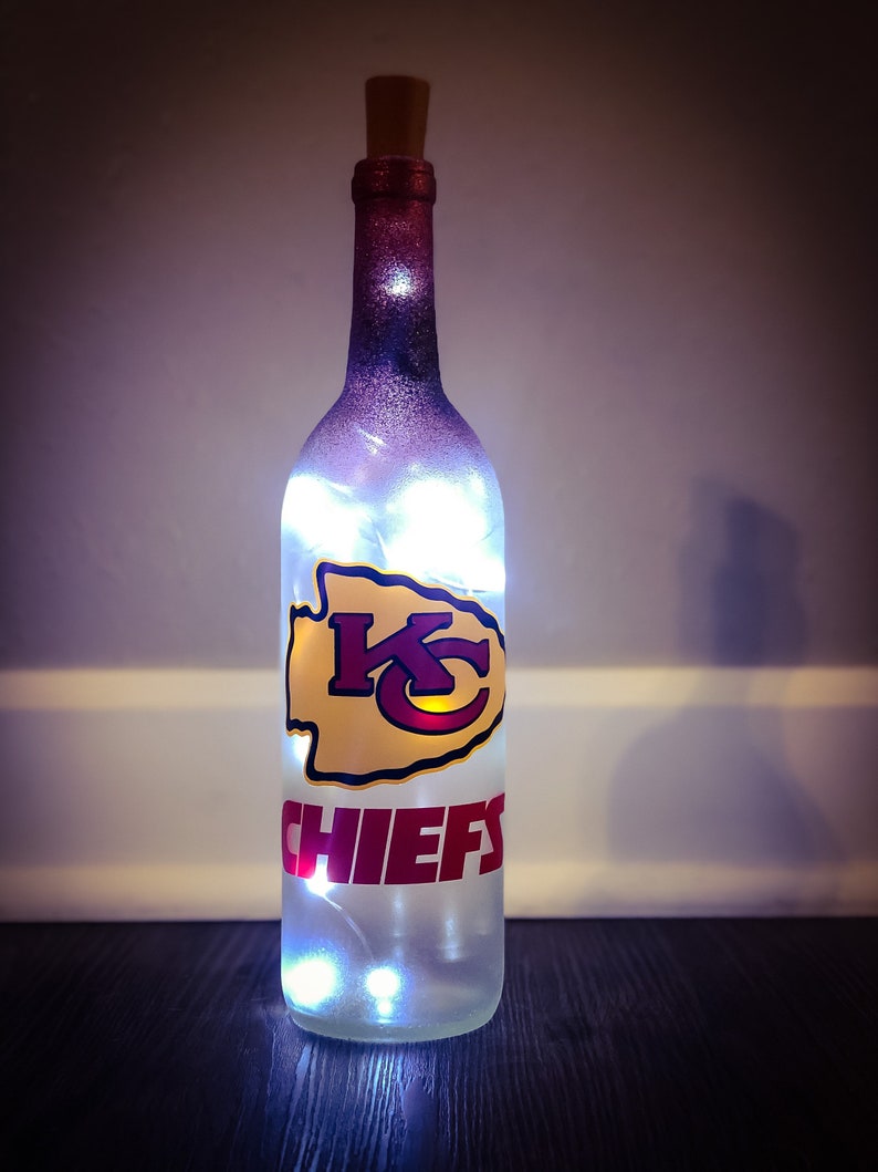 May include: A clear glass bottle with a white and purple ombre paint finish. The bottle has a cork stopper and features a red and yellow Kansas City Chiefs logo with the word "CHIEFS" in red lettering. The bottle is lit with white fairy lights.