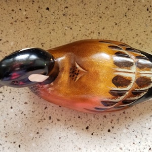May include: A wooden duck decoy with brown, black, and white painted details. The duck has a black head and a white patch on its cheek. The body is painted with brown and white feathers.
