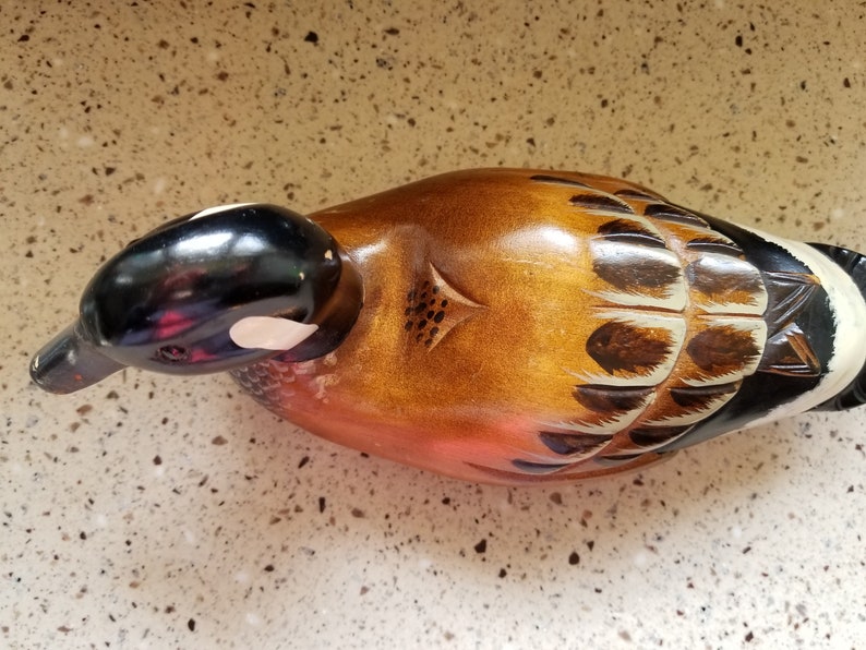 May include: A wooden duck decoy with brown, black, and white painted details. The duck has a black head and a white patch on its cheek. The body is painted with brown and white feathers.