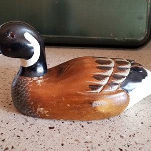 May include: A wooden carved duck decoy with brown, black, and white paint. The duck has a long black beak and a white ring around its neck.