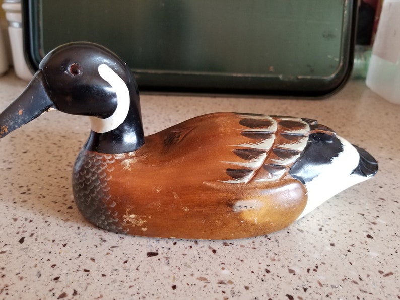 May include: A wooden carved duck decoy with brown, black, and white paint. The duck has a long black beak and a white ring around its neck.
