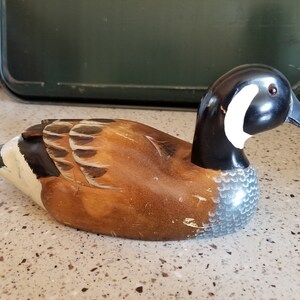 May include: A wooden carved duck decoy with brown, black, white, and gray paint. The duck has a long neck and a detailed head with a black beak and a white stripe.