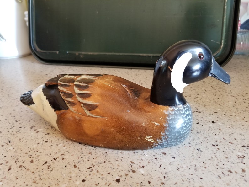 May include: A wooden carved duck decoy with brown, black, white, and gray paint. The duck has a long neck and a detailed head with a black beak and a white stripe.