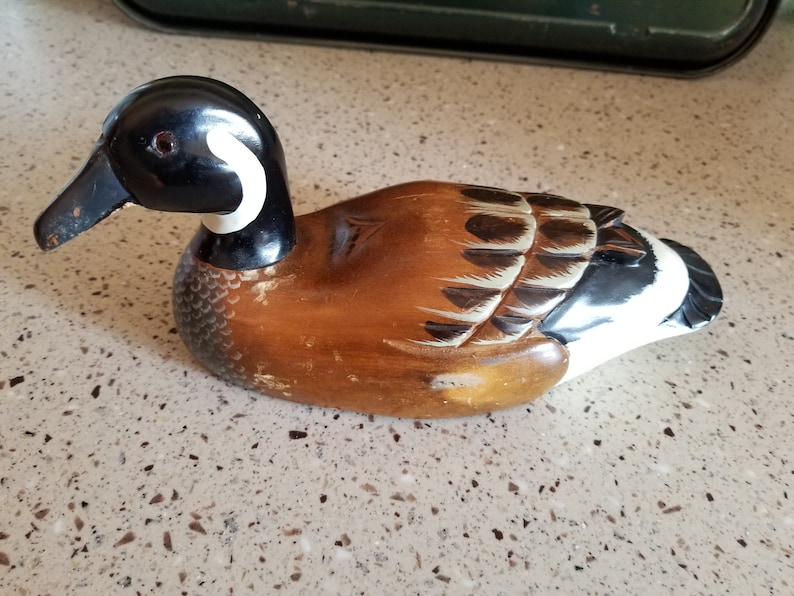 May include: A wooden carved duck decoy with brown, black, and white paint. The duck has a black head with a white stripe around the neck. The body is brown with black and white markings.