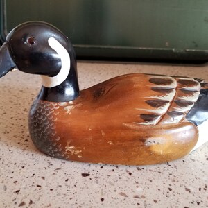 May include: A wooden carved duck decoy with brown, black, and white paint. The duck has a black head with a white ring around the neck. The body is brown with black and white markings.