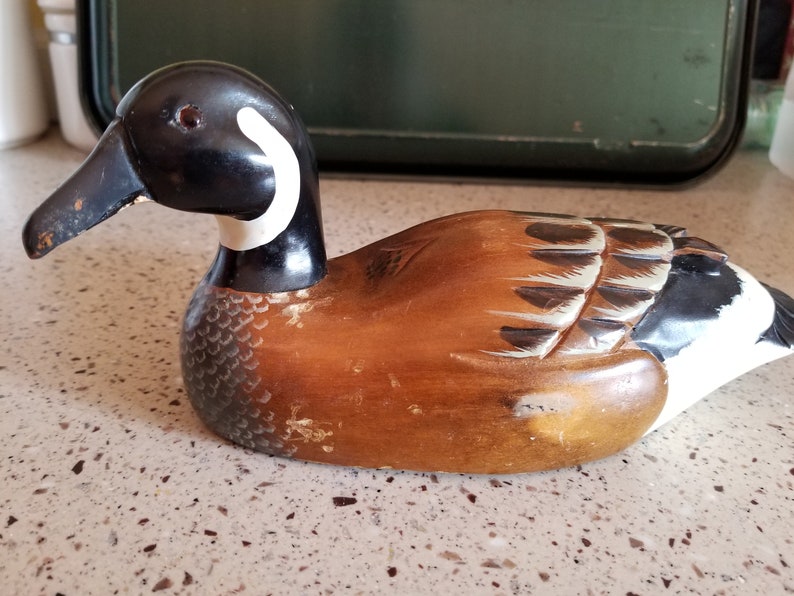 May include: A wooden carved duck decoy with brown, black, and white paint. The duck has a black head with a white ring around the neck. The body is brown with black and white markings.