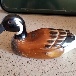May include: Hand-carved wooden duck decoy with brown, black, and white paint. The duck has a black head with a white stripe and a brown body with black and white wing markings.