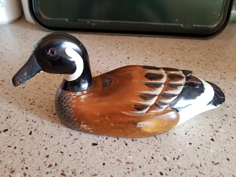 May include: Hand-carved wooden duck decoy with brown, black, and white paint. The duck has a black head with a white stripe and a brown body with black and white wing markings.